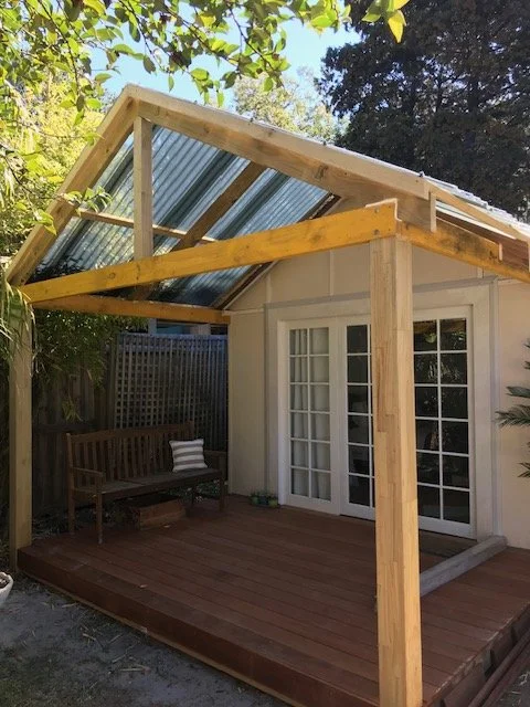 Outdoor backyard structure with a wooden deck, a small building with glass doors, and an unfinished roof frame with corrugated metal roofing panels.