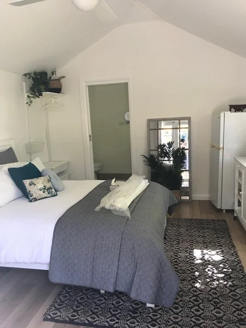 A cozy bedroom with a white bed, dark gray blanket, decorative pillows, and a small white shelf with potted plant, in a room with white walls and wooden floor.