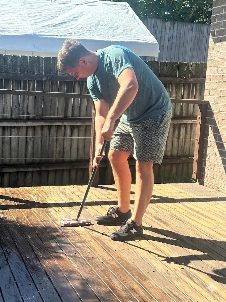 A man cleaning a wooden deck outdoors with a mop on a sunny day.