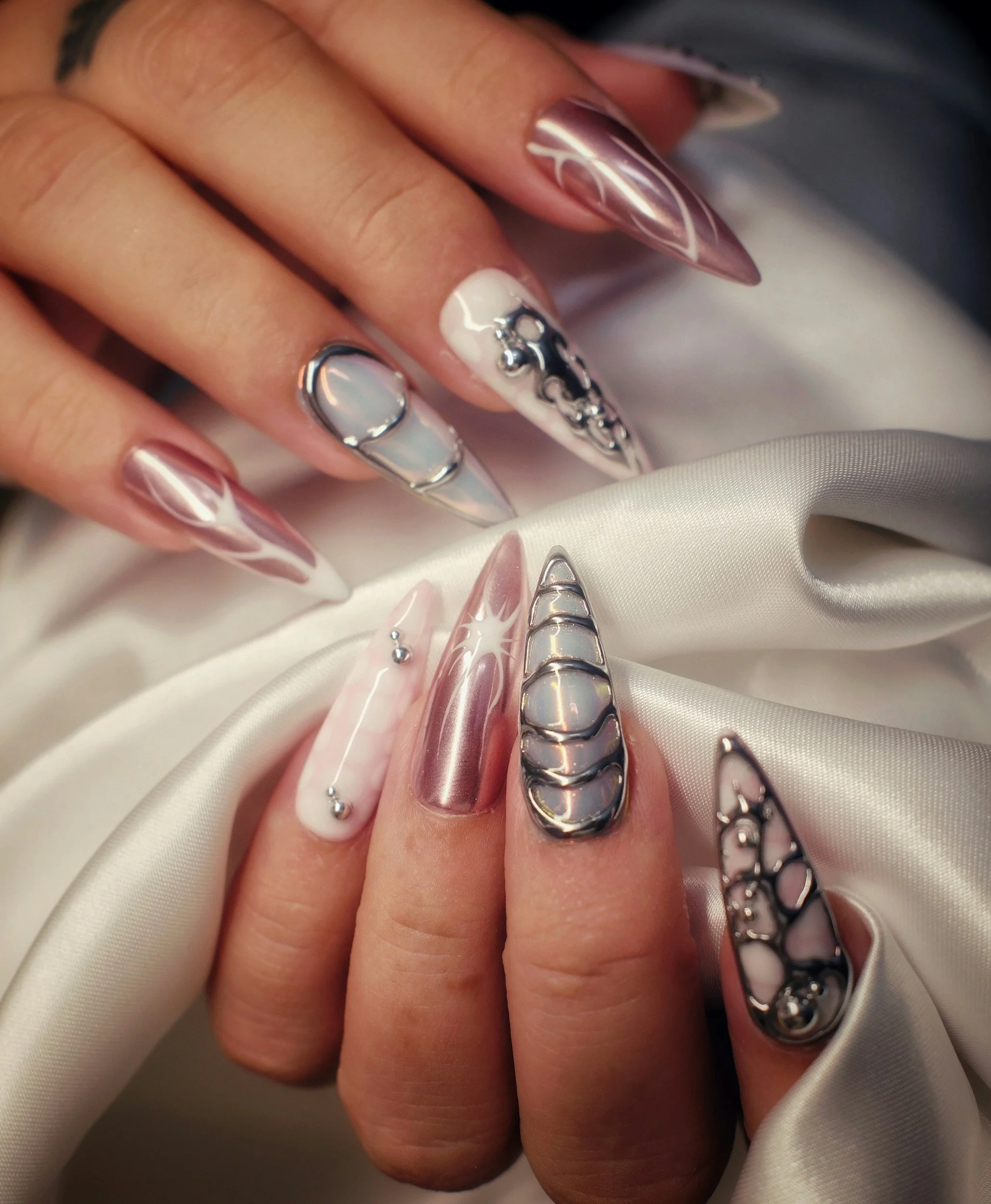 Close-up of a hand with long, almond-shaped nails featuring a marble design and chrome accents and trending nail art, resting on a silky fabric.