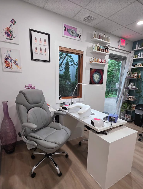 Interior of Nail Network with a white desk, gray ergonomic chair, nail equipment, wall art, shelves with nail polish bottles, window showing outdoor greenery, and door with an exit sign.