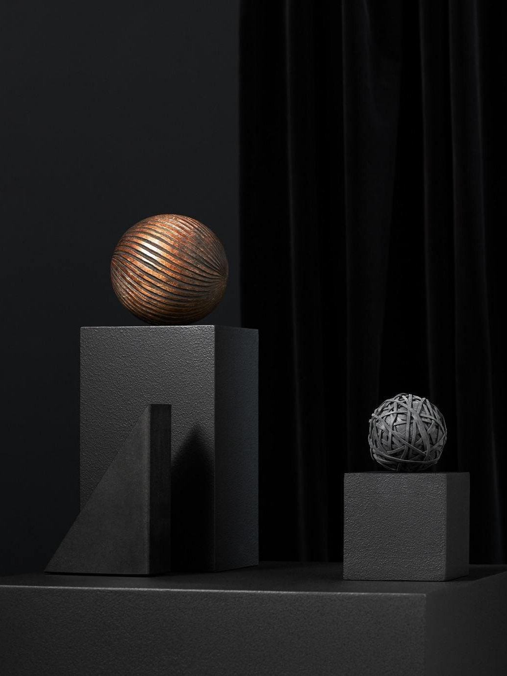 Two decorative spheres on textured pedestals; one metallic with bronze finish and stripes, the other gray with a tangled design, set against a dark background with black curtains.