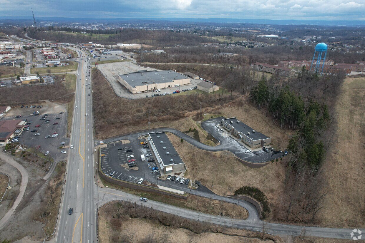 An aerial view of a commercial area with parking lots, a water tower, a large building, and surrounding roads and terrain.