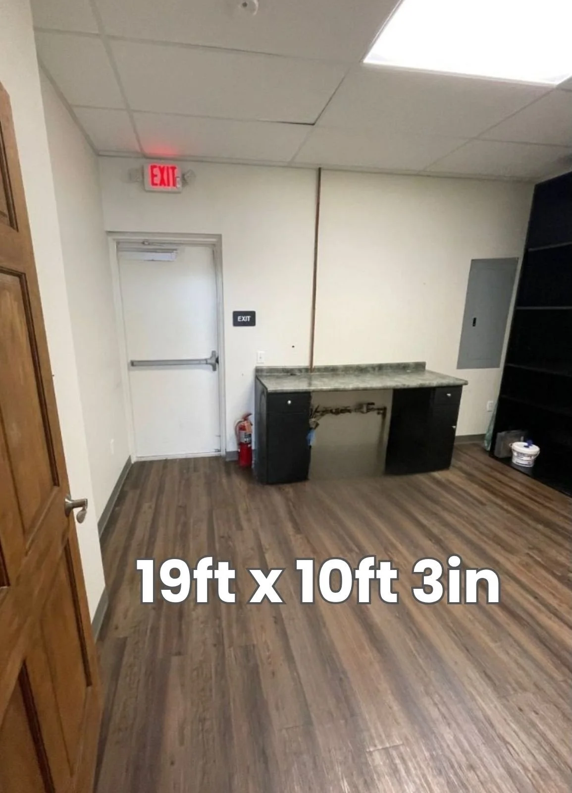 Empty room with wooden flooring, a white door with an exit sign above, a small black desk with a marble top, a fire extinguisher nearby, and built-in shelves on the right. The room dimensions are 19 feet by 10 feet 3 inches.