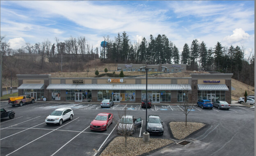A strip mall with multiple storefronts, a parking lot with several parked cars, and a small hill with trees and a water tower in the background.