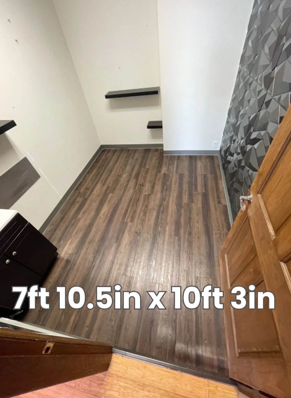 Empty room with wood flooring, white and patterned accent walls, black floating shelves, and wooden door.