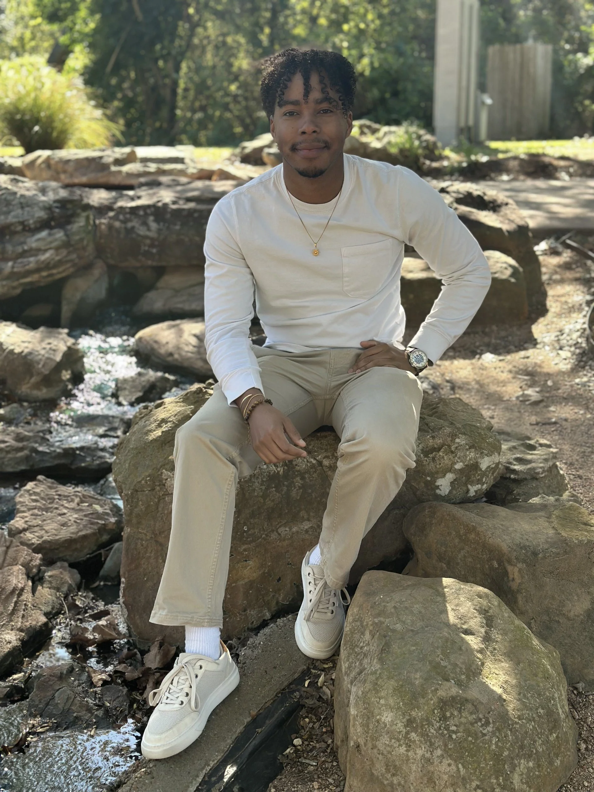 Young man sitting on rocks beside a small stream, outdoors, wearing a white long-sleeve shirt, beige pants, white sneakers, and a watch, with trees and a wooden fence in the background.