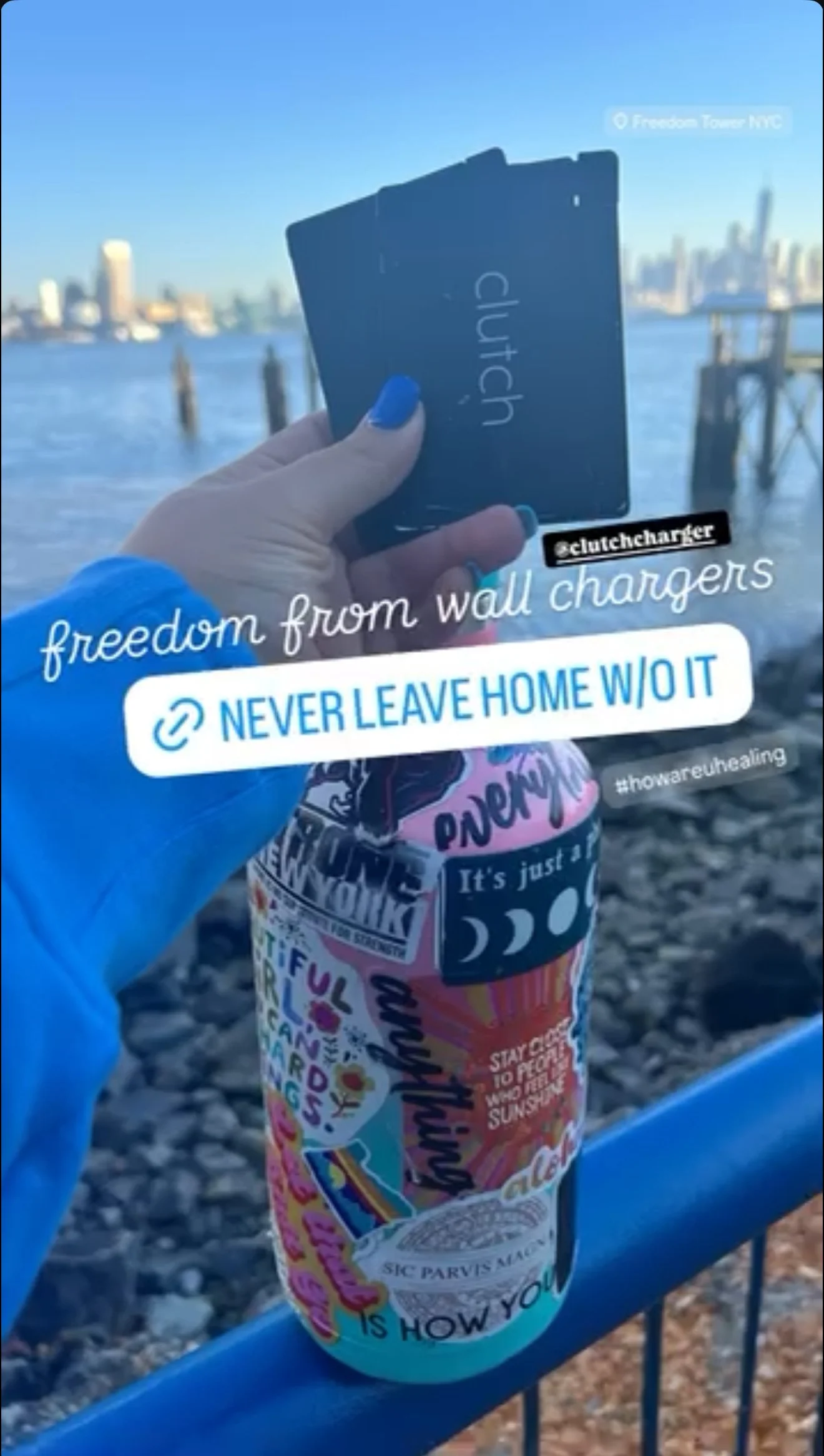 Person with blue nails holding "clutch" branded cards near a sticker-covered bottle by the waterfront with a cityscape background.