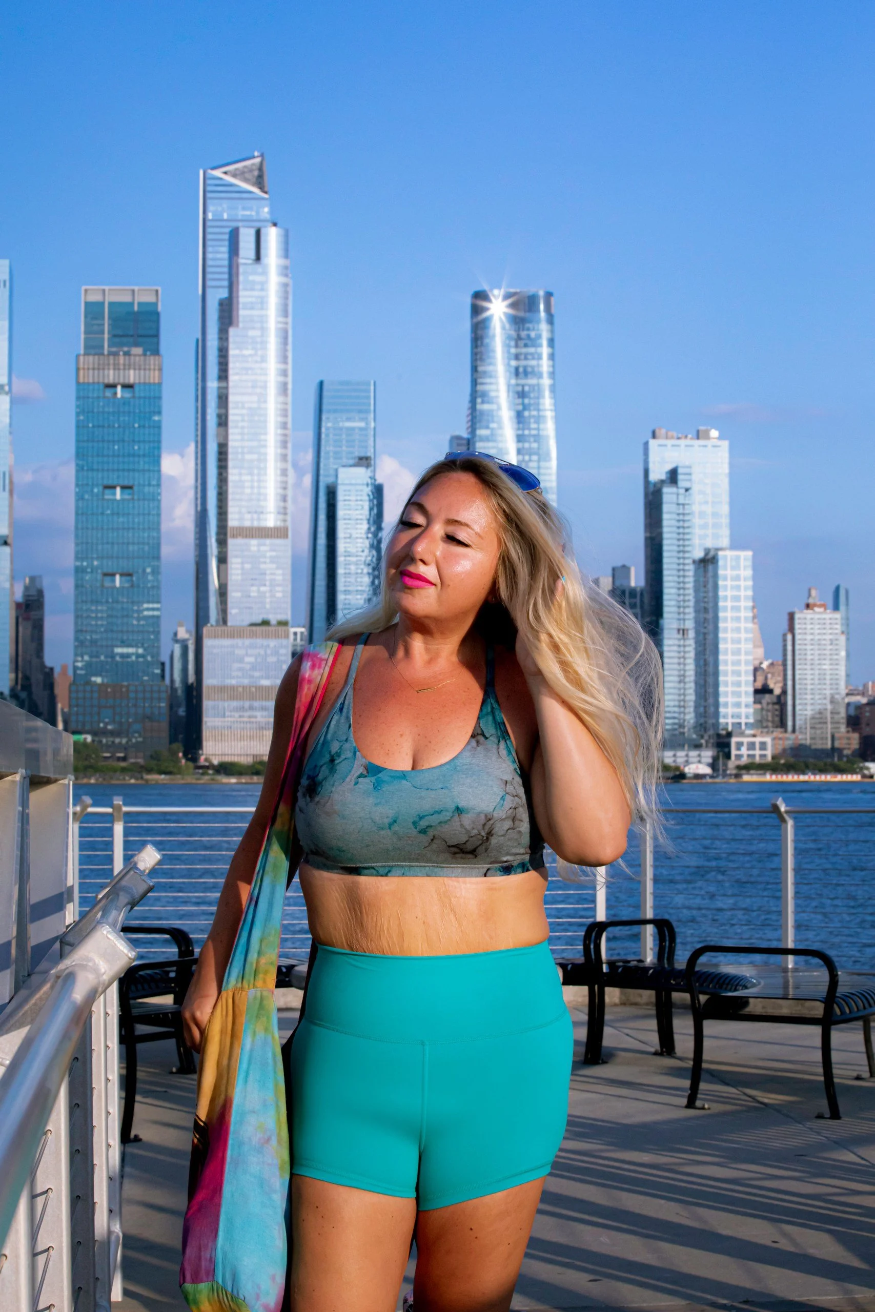 Woman in workout attire with blonde hair stands on a waterfront pathway with New York City skyline in the background.