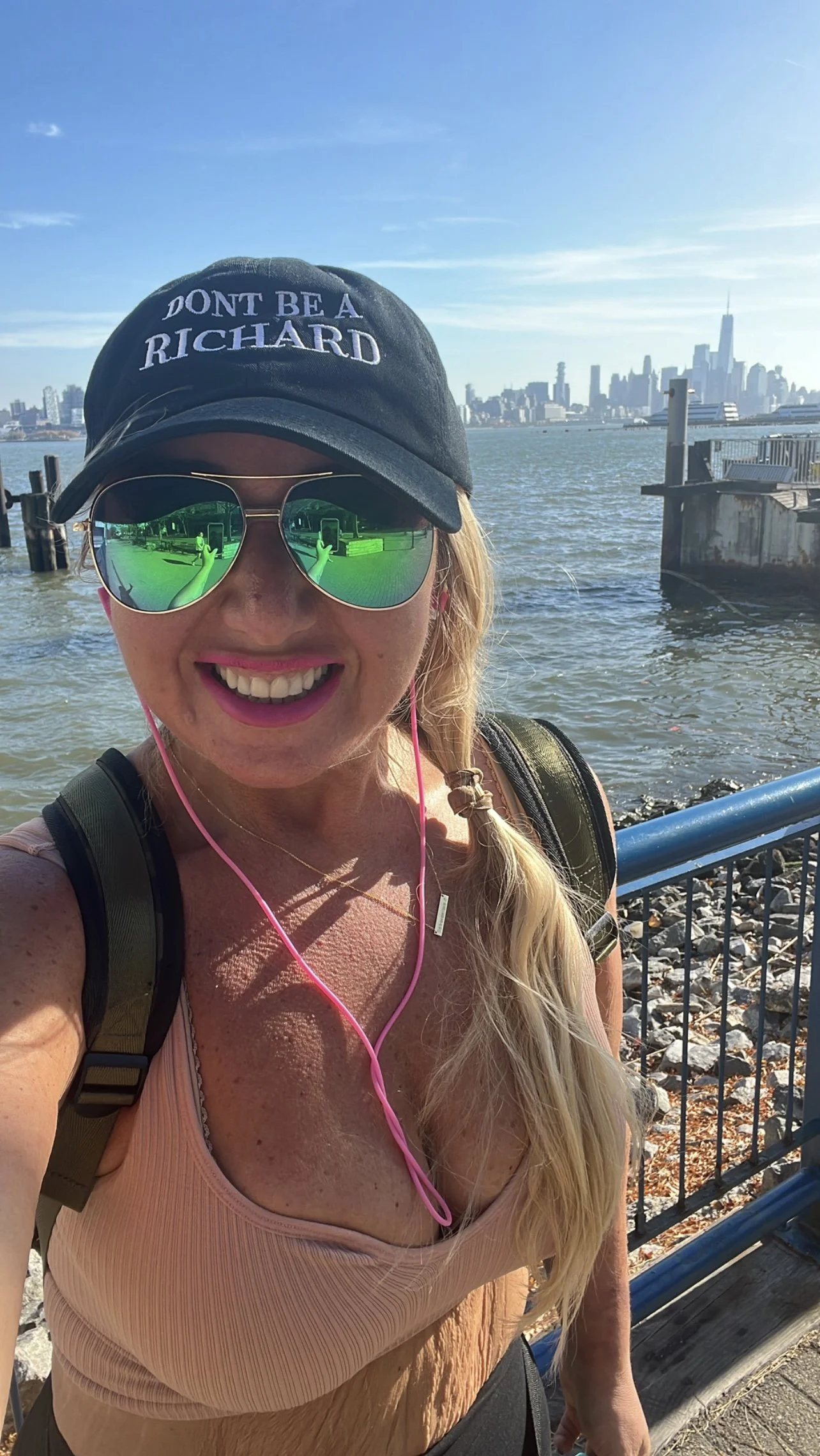 Lauren wearing a hat with "DON'T BE A RICHARD" text, sunglasses, and earphones, standing near water with a city skyline in the background.