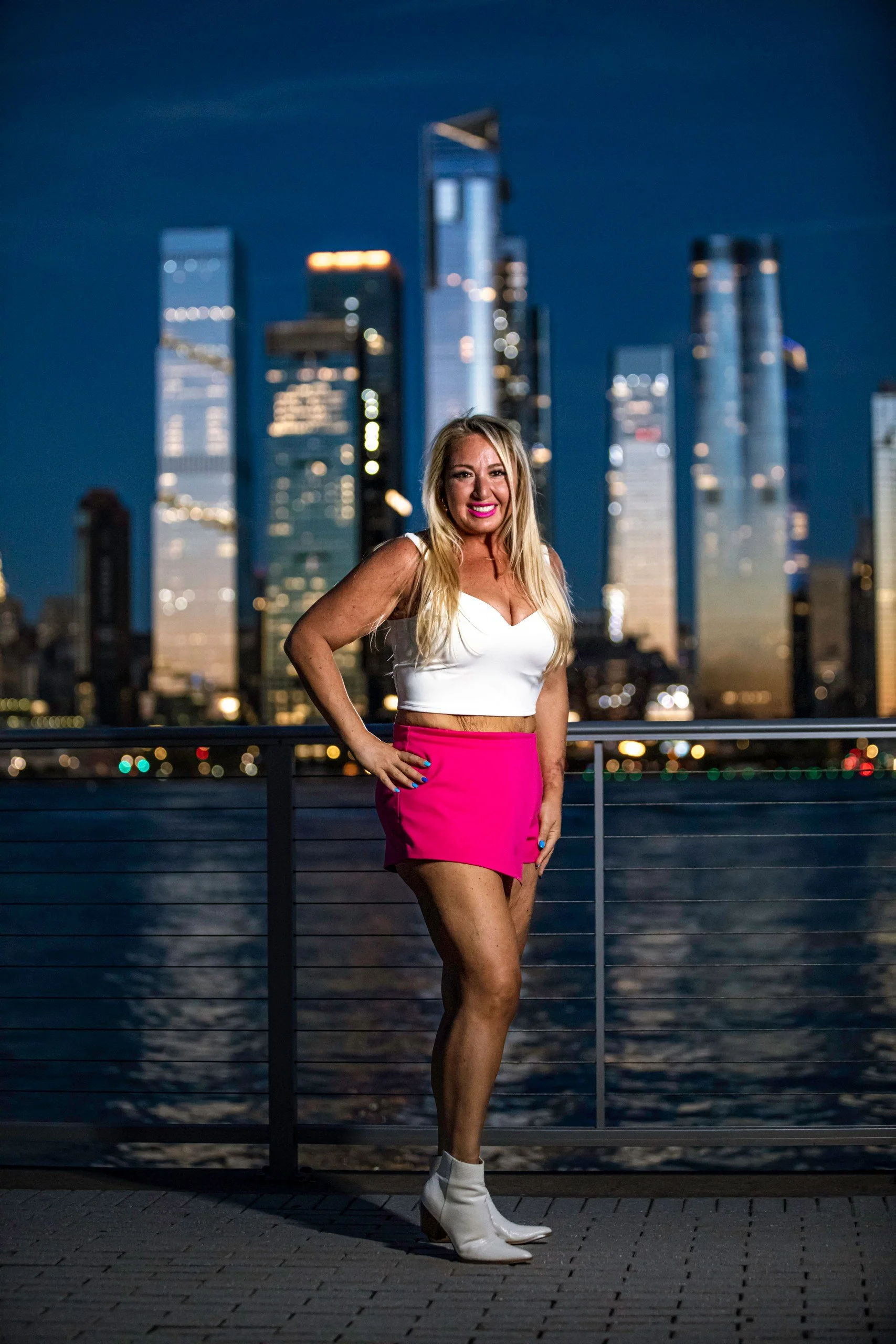 Person posing on a waterfront with city skyscrapers in the background at night.