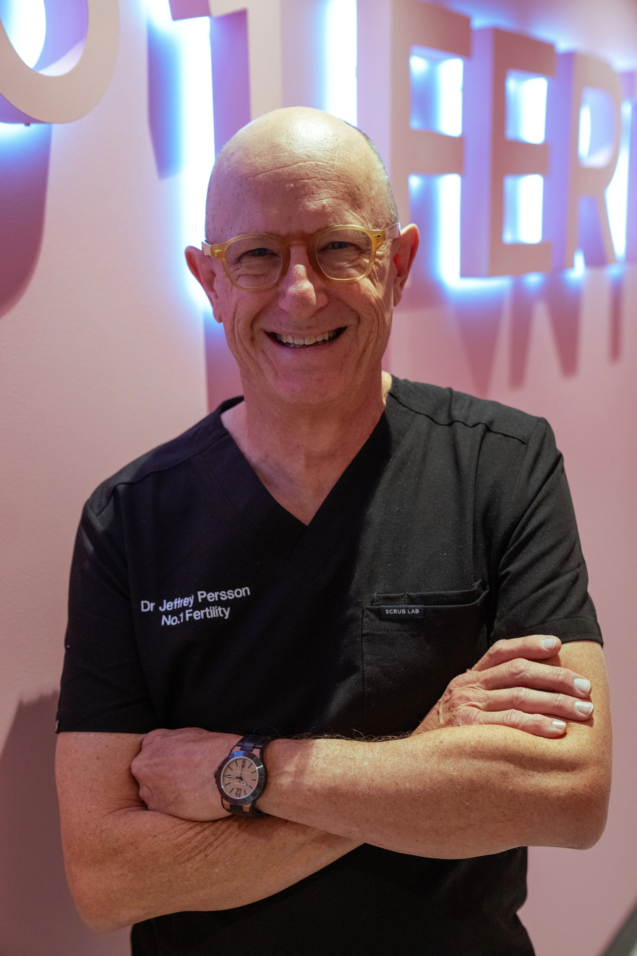 A smiling man - Dr Jeffrey Persson - wearing a black medical uniform with text on it, standing with arms crossed in front of a pink wall with backlit decor spelling out 'No 1 Fertility'.
