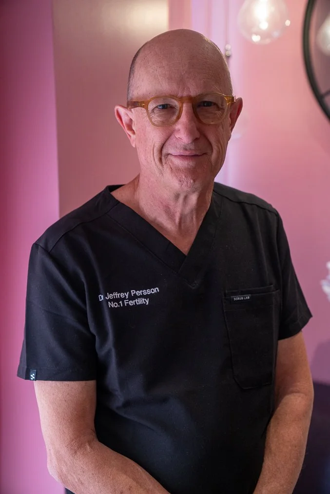 Dr Jeffrey Person wearing glasses and a black medical scrub top with the name 'Dr Jeffrey Persson' and the phrase 'No.1 Fertility' embroidered on it, standing in a room with pink walls and a round mirror.