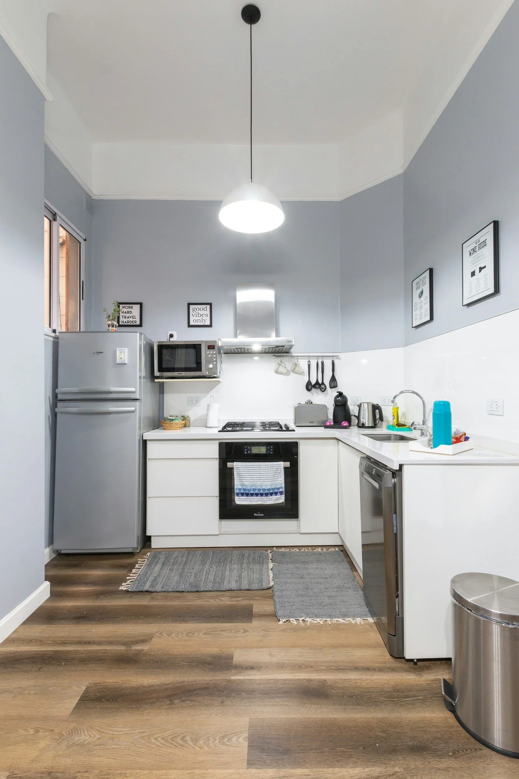 Modern kitchen with white cabinets, stainless steel refrigerator, microwave, and oven. Gray walls with framed quotes and posters. Items on the countertop include a toaster, kettle, utensils, and cleaning supplies. Wooden flooring with gray rugs.