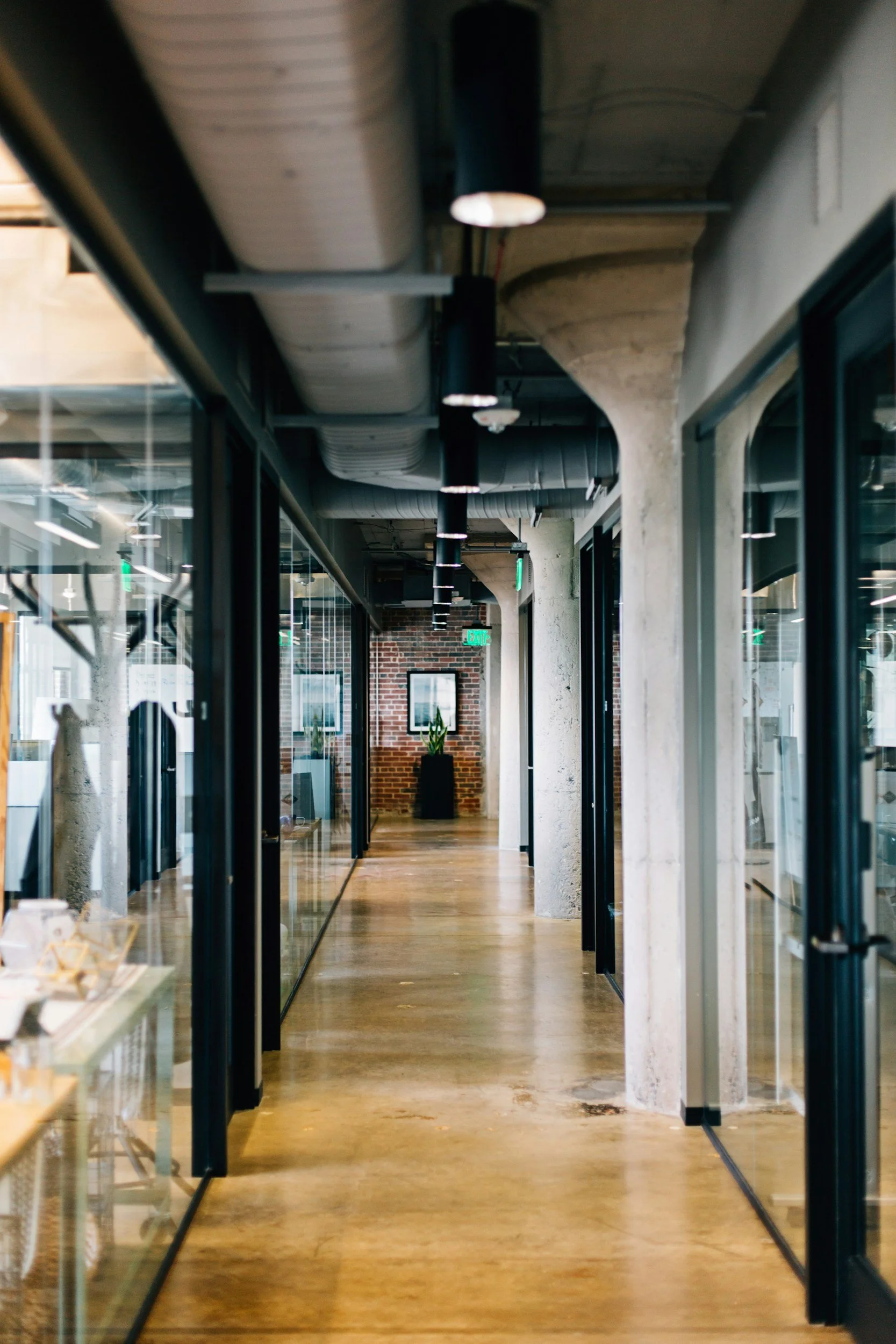 A modern office corridor with glass walls, open ceiling with visible ductwork and black pendant lights, wooden flooring, and potted plants at the end of the hallway.