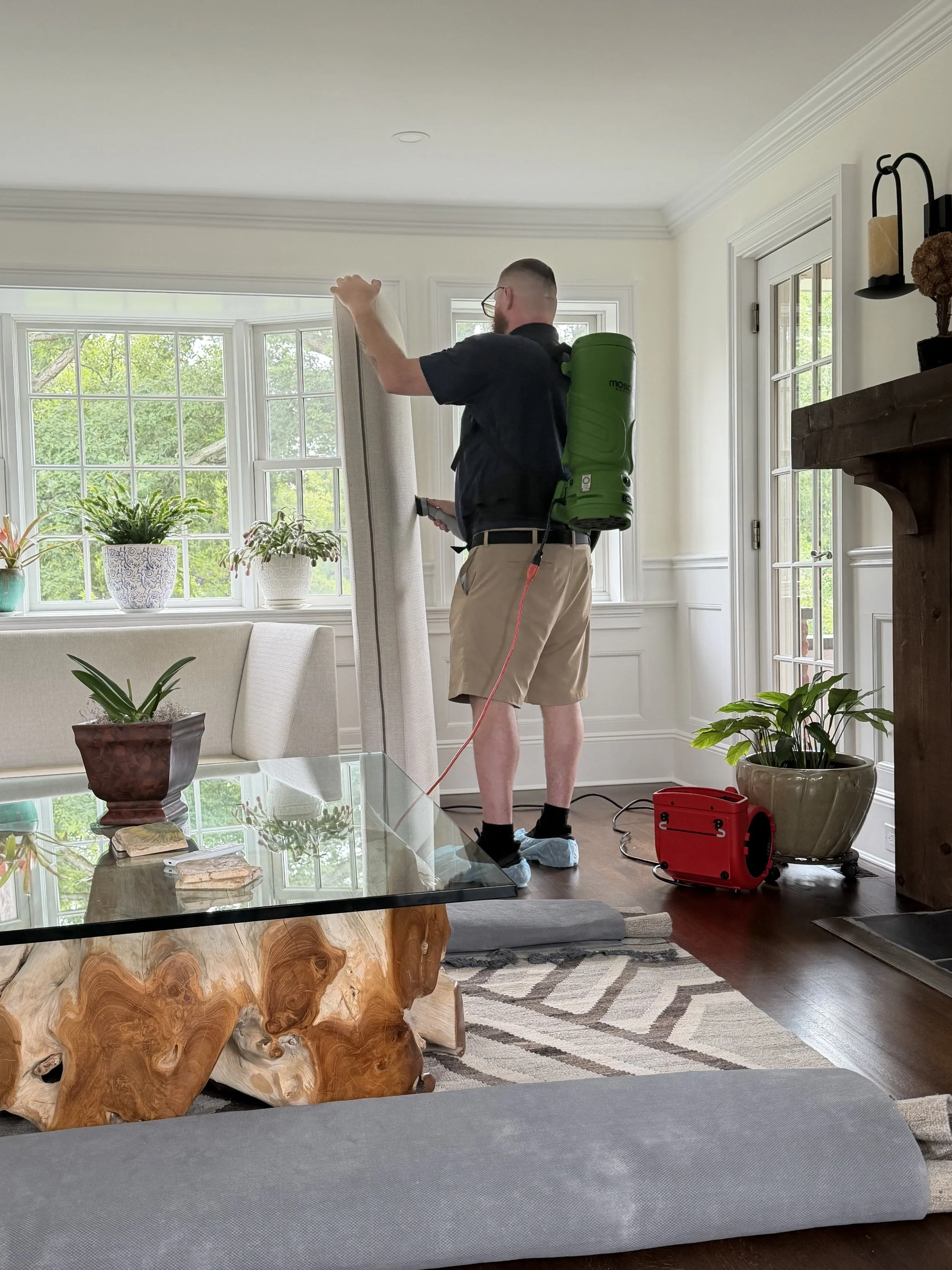 Man in black shirt and khaki shorts cleaning a window in a bright living room, carrying a robotic vacuum on his back, with plants and furniture visible.