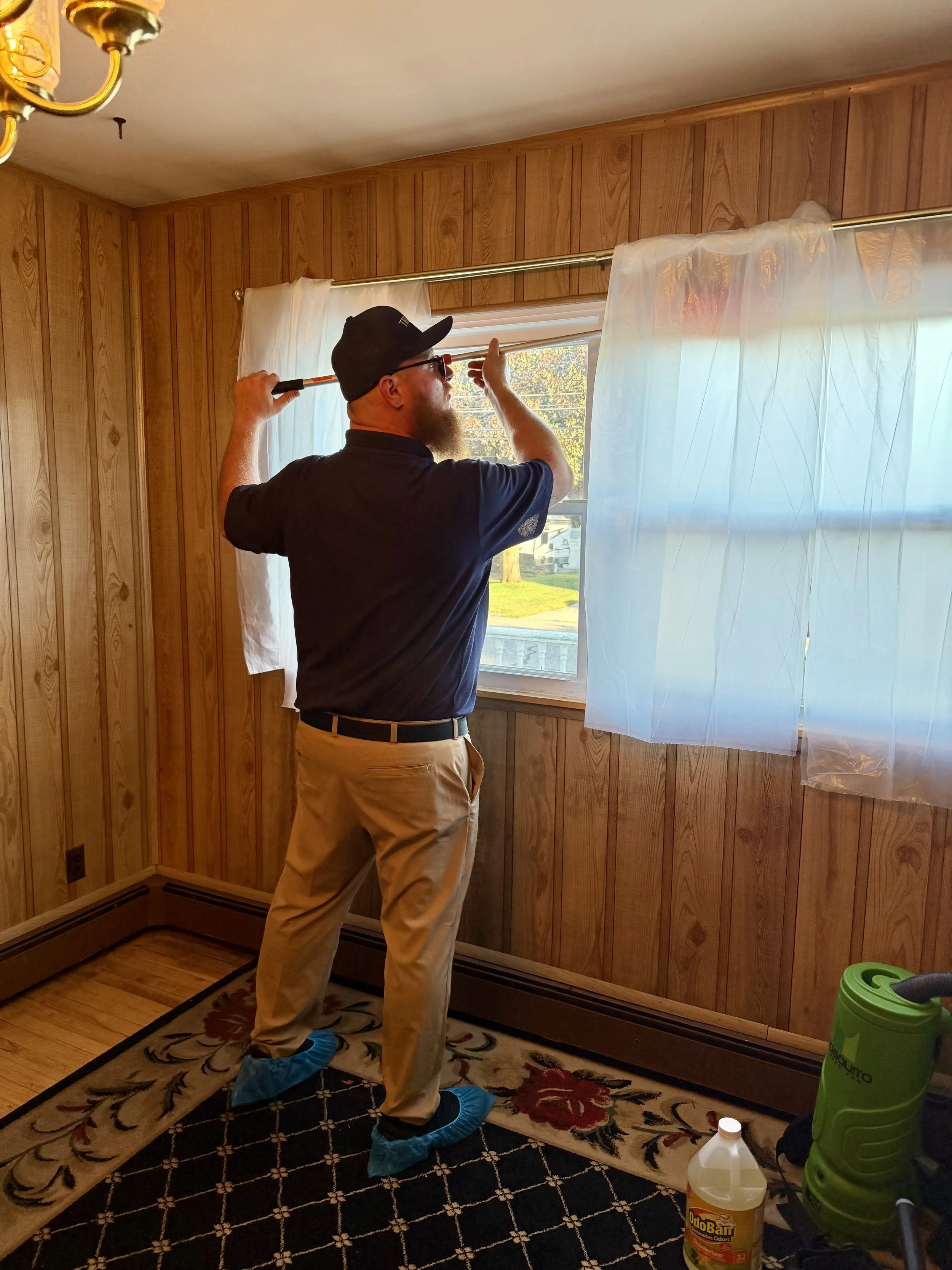 A man wearing a black cap, sunglasses, and blue shoe covers is opening a window in a room with wooden panel walls. There are cleaning supplies on the floor, including a yellow OdorBan bottle, a green vacuum, and a white spray bottle.