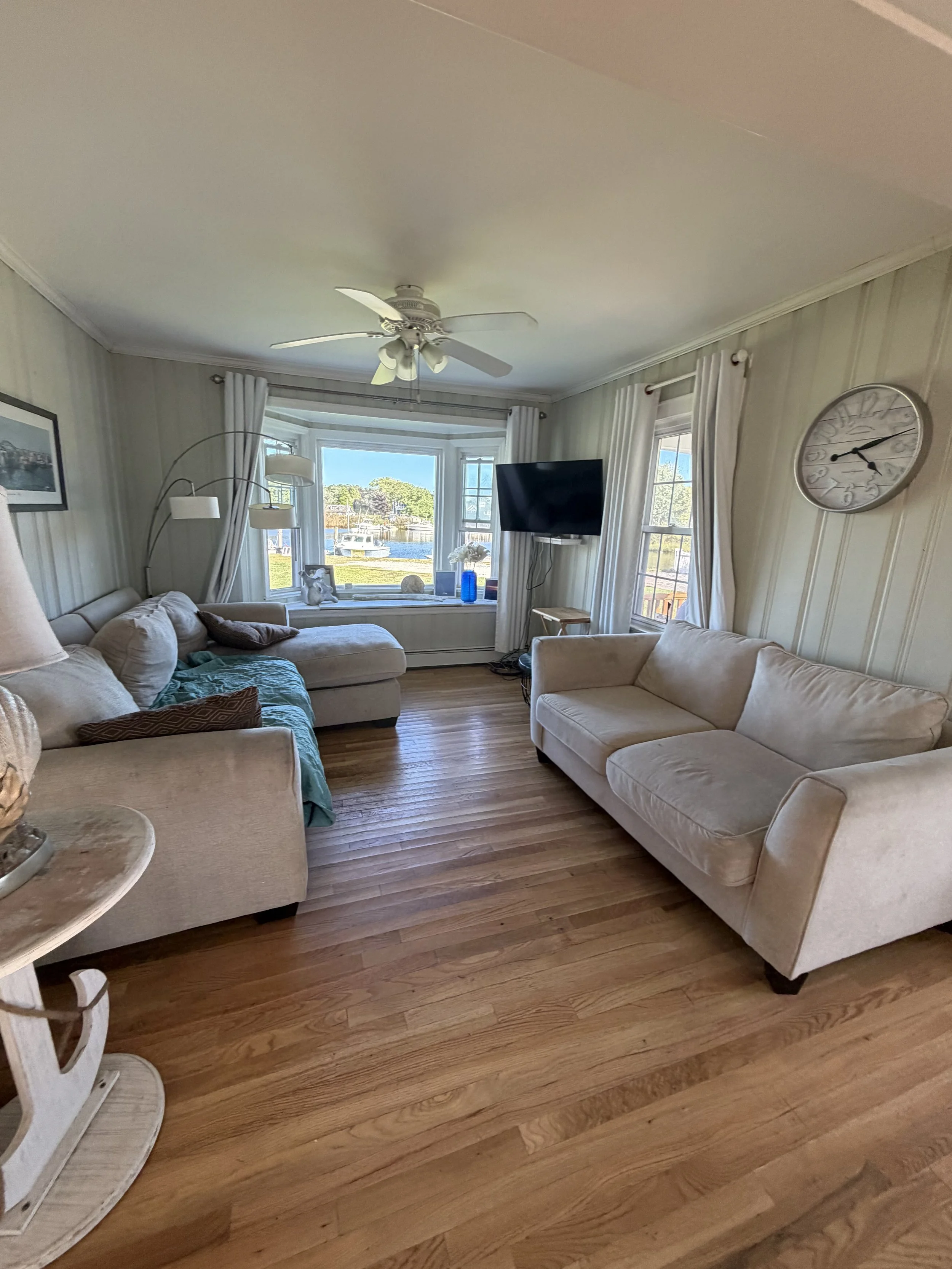 Living room with beige couches, hardwood floor, windows with white curtains, a wall clock, a TV, a standing lamp, and a view of boats and water outside.