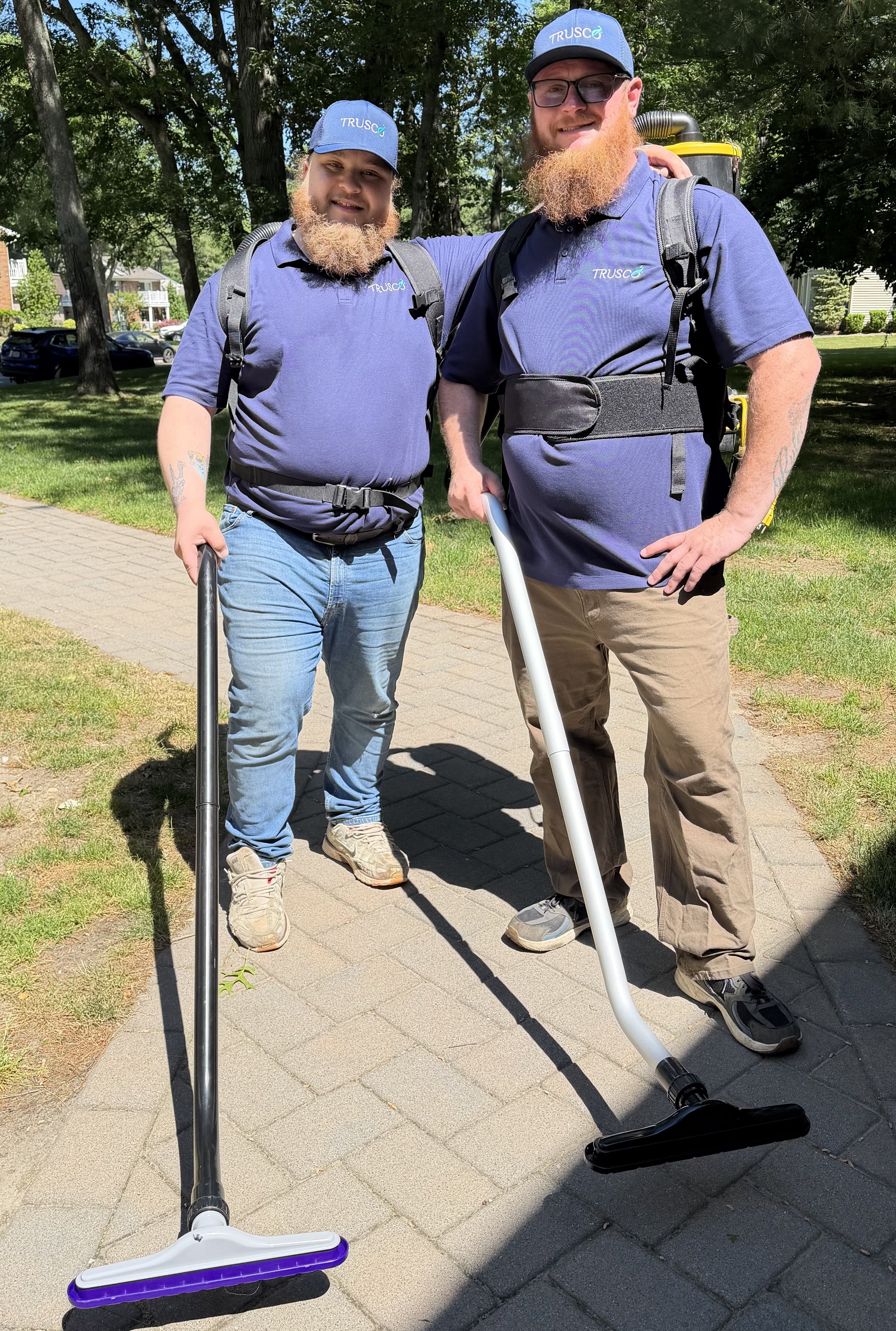 Two men with ginger beards wearing blue Trusco uniforms, caps, and backpacks, standing outdoors on a sidewalk with trees and houses in the background, each holding a cordless vacuum cleaner.
