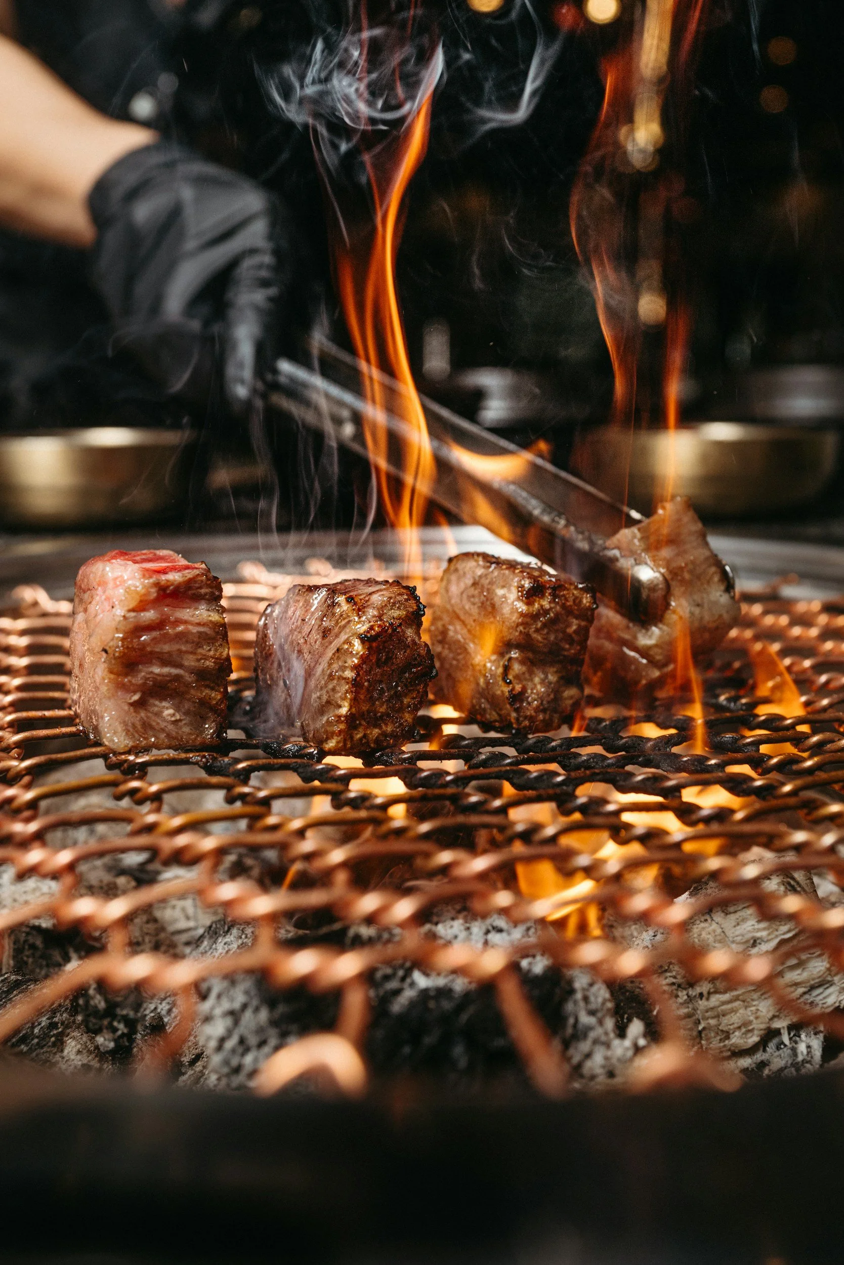 Close-up of sizzling meat on a grill with flames and smoke rising, with a hand using tongs in the background.