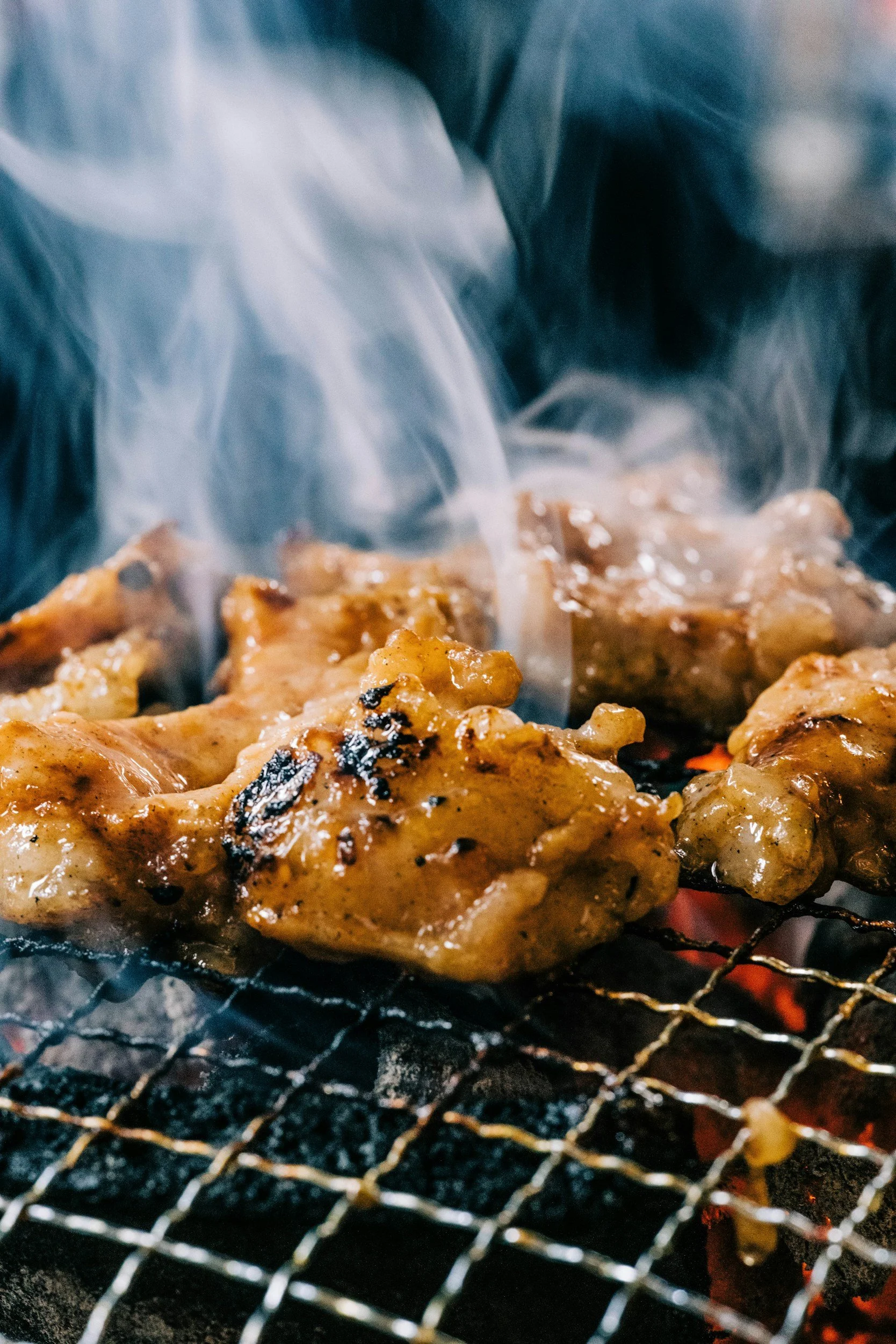 Close-up of chicken pieces grilling on a barbecue, with smoke rising and glowing coals underneath.