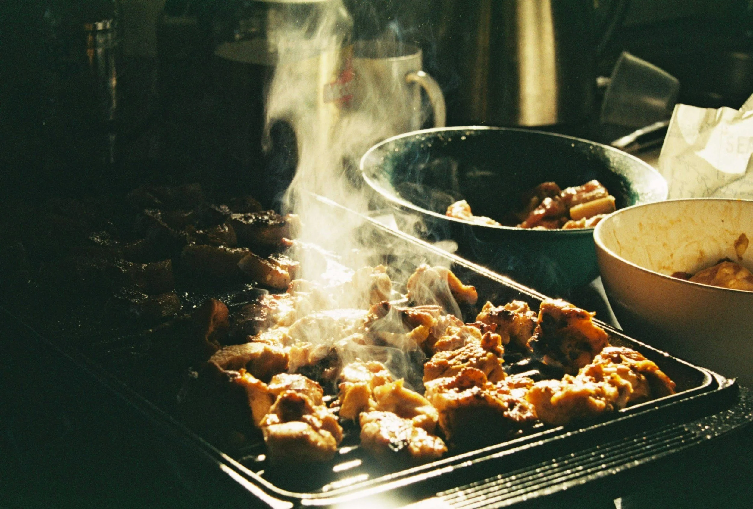Meat grilling on an open flame grill with smoke rising, with bowls nearby in a kitchen setting.