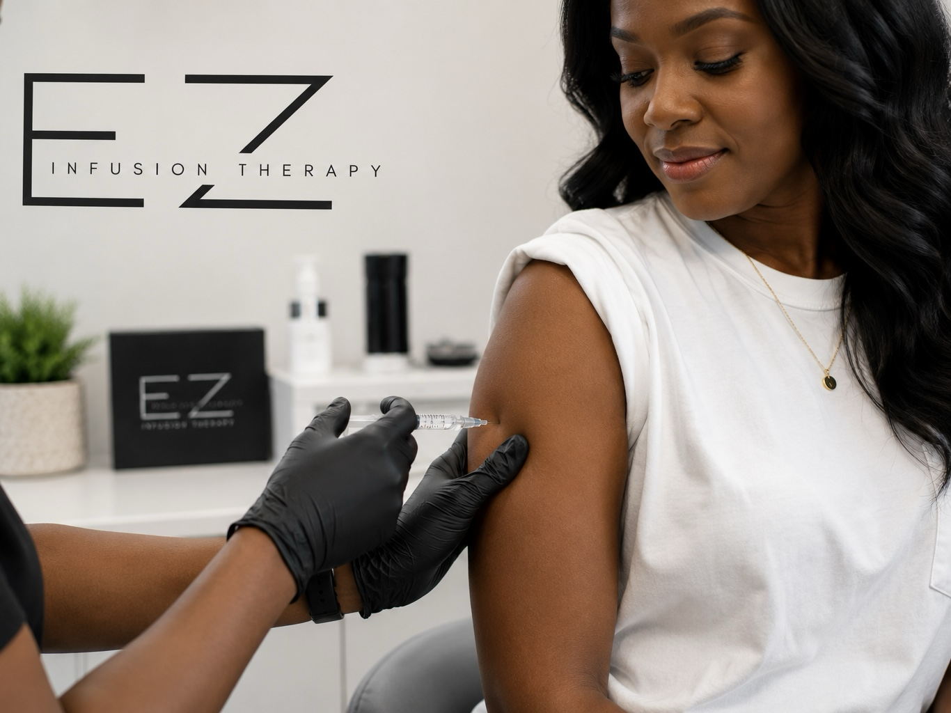 A woman receiving a vaccination in her upper arm from a healthcare professional wearing black gloves, in a medical office with a sign for EZ Infusion Therapy.