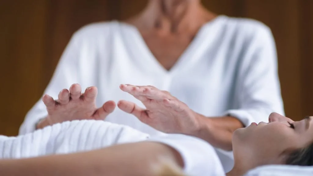 A person lying down with their eyes closed, while another person appears to be administering some form of healing or therapy using their hands.