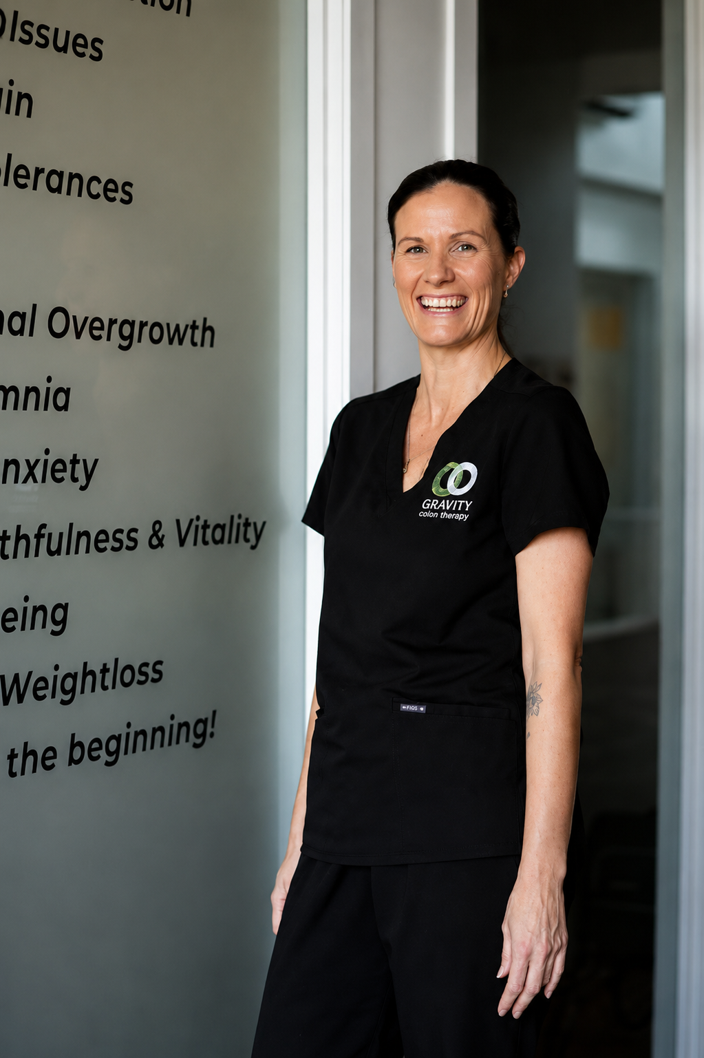 A woman in black scrubs smiling and standing indoors next to a list of health-related issues written on a gray wall.
