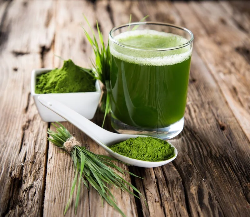 A glass of bright green smoothie, a bowl of green powder, and a spoonful of green powder on a white ceramic spoon resting on a rustic wooden surface, with a sprig of green grass nearby.