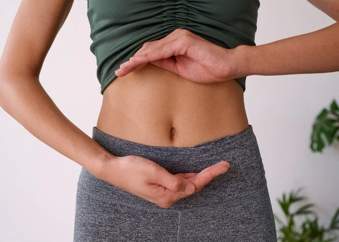 Close-up of a woman's midsection showing her wearing a green top and gray leggings, with her hand forming a heart shape around her stomach.
