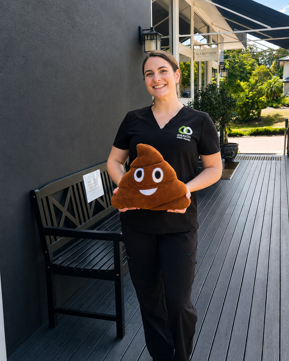 A woman in black scrubs standing outdoors, smiling, holding a plush toy shaped like a smiling pile of poop with cartoon eyes and mouth.