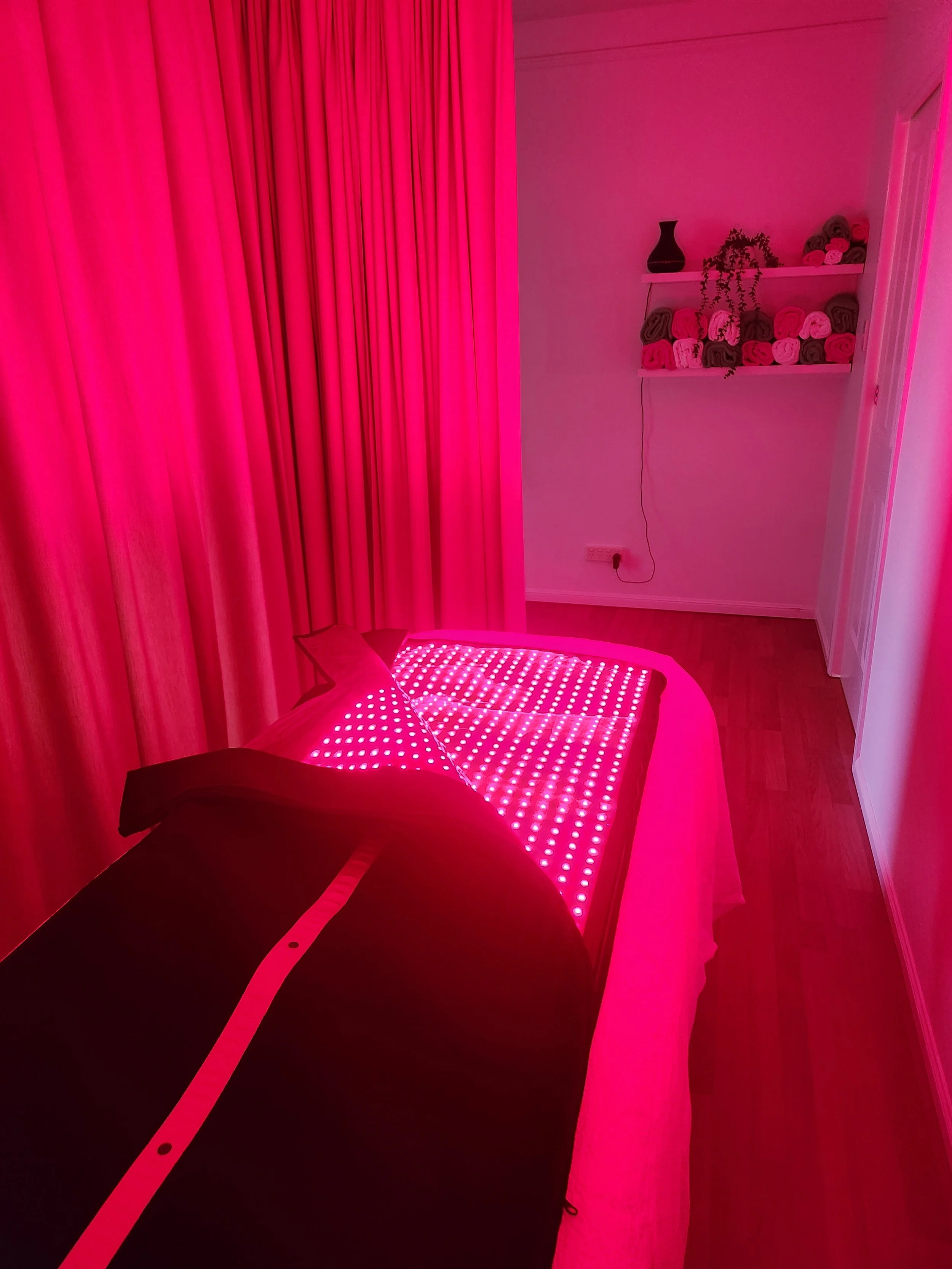 A room illuminated with pink lighting, featuring a black chair, a pink curved surface with LED lights, pink curtains, and white shelves with rolled towels, a black vase, and a small plant.