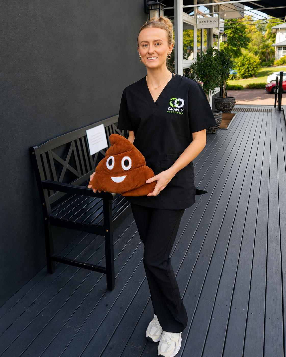 A woman standing outdoors on a wooden deck, holding a plush toy shaped like a smiling poop emoji. She is wearing black scrubs with a logo that reads 'GRAVITY colon therapy.'