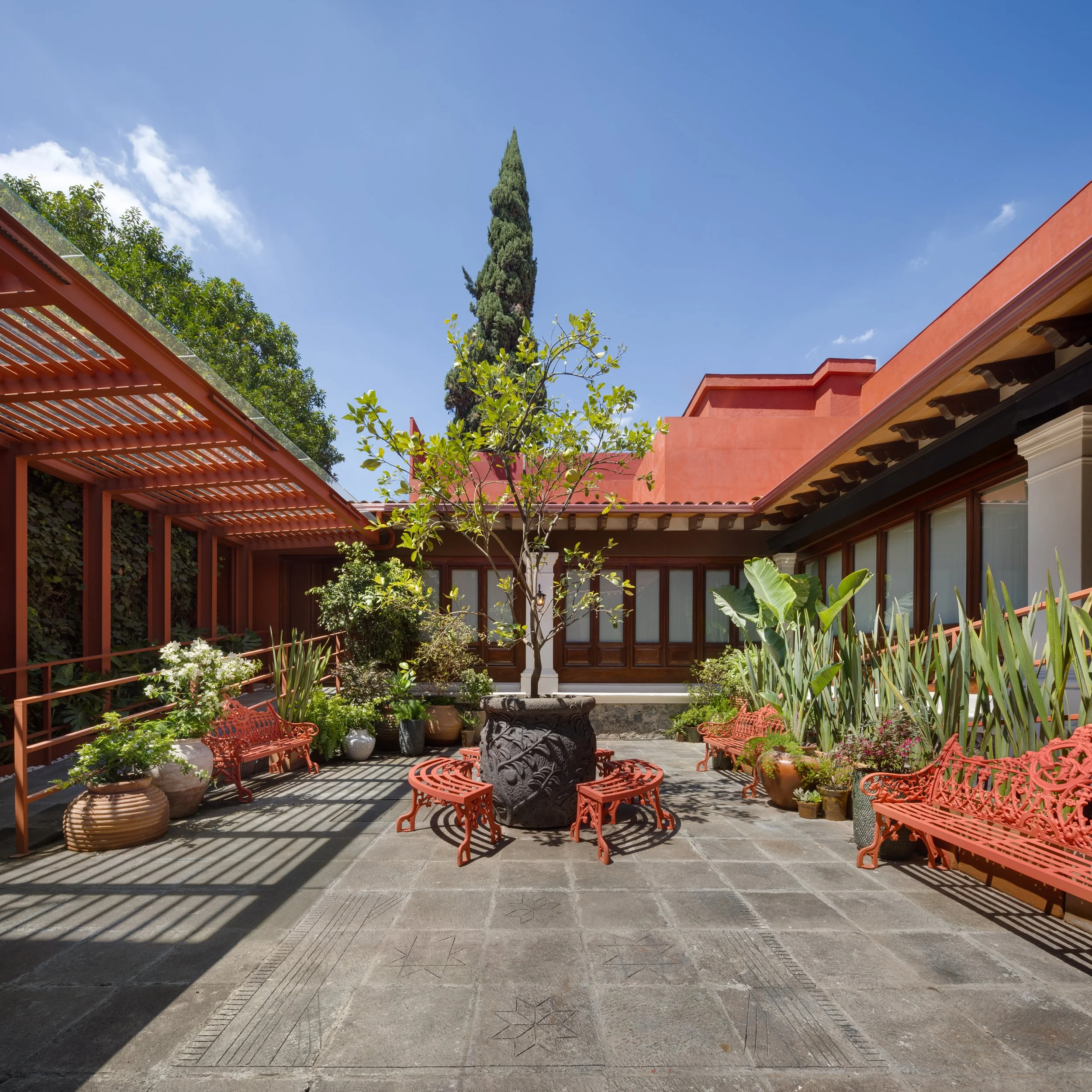 A courtyard garden with red metal chairs and benches, potted plants, and a courtyard fountain, surrounded by red and beige buildings with arched windows and rooftop, under a clear blue sky with some clouds.