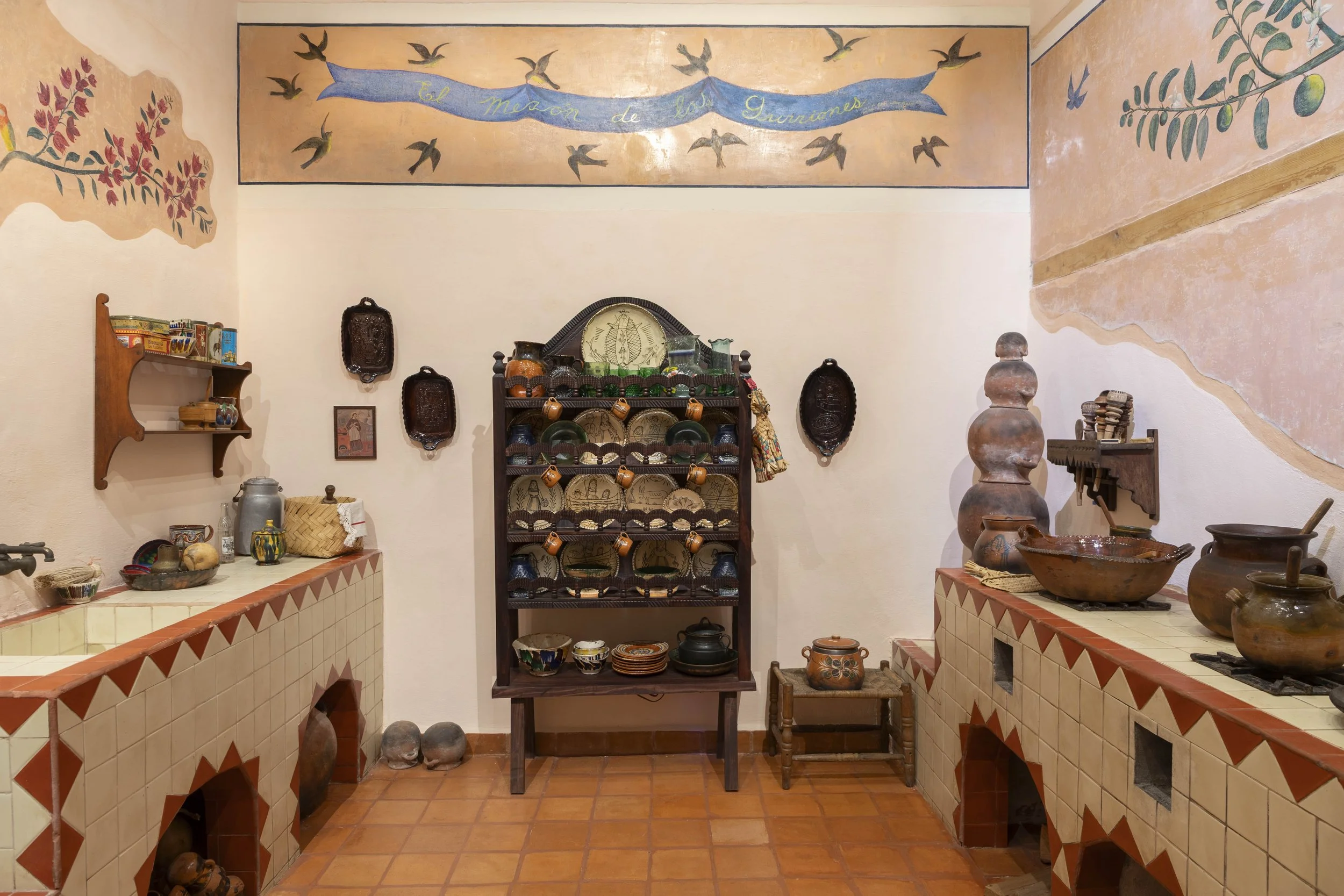 Interior of a rustic kitchen or display area with wooden shelves, pottery, and decorative items. There are painted walls with a mural of birds and a banner reading 'El Meson de Los Guardianes' at the top.