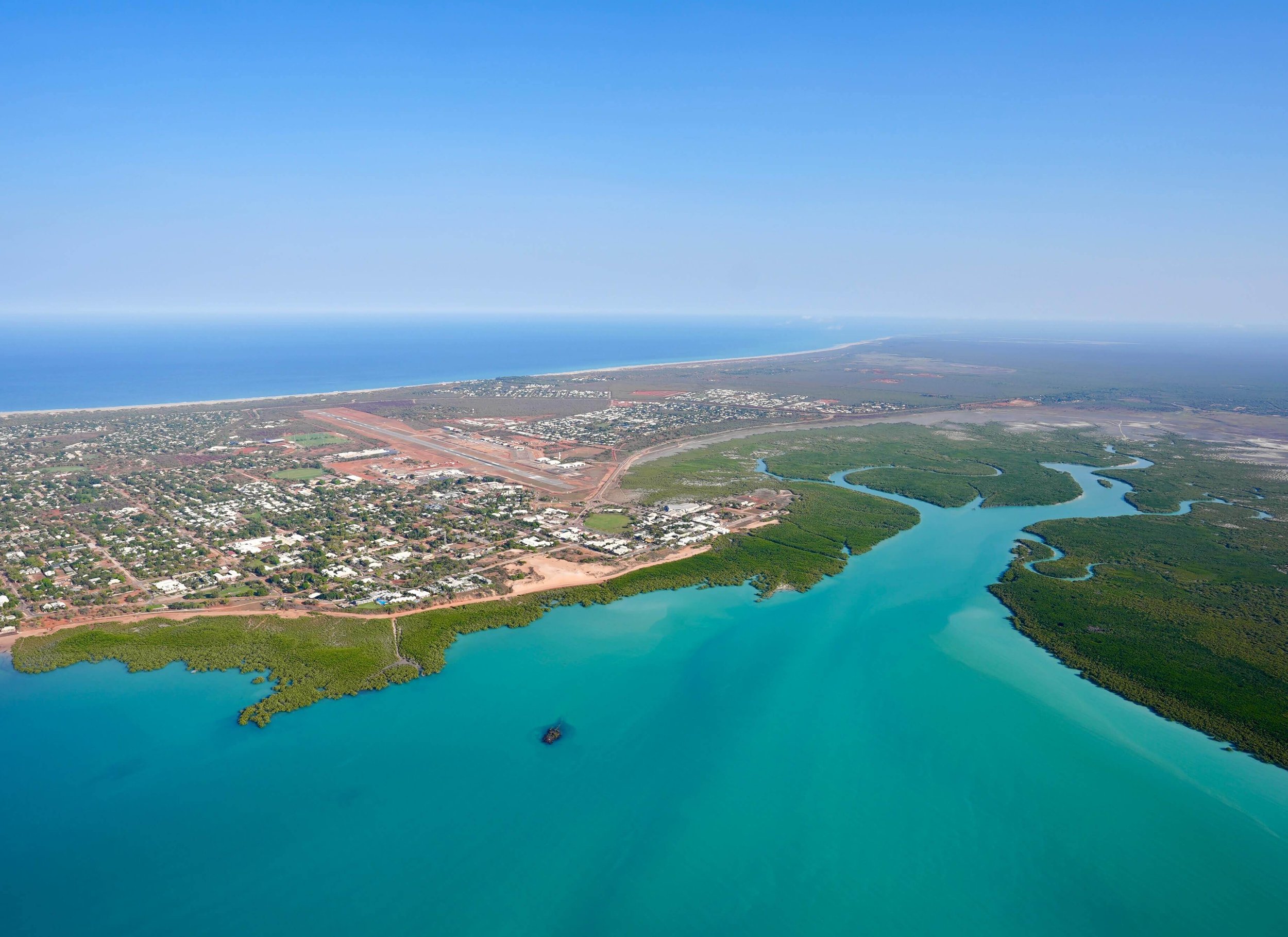 An aerial view of a coastal area showing a town near the shoreline, a large body of water with a small boat, green mangrove forests, and the ocean with a clear blue sky above.