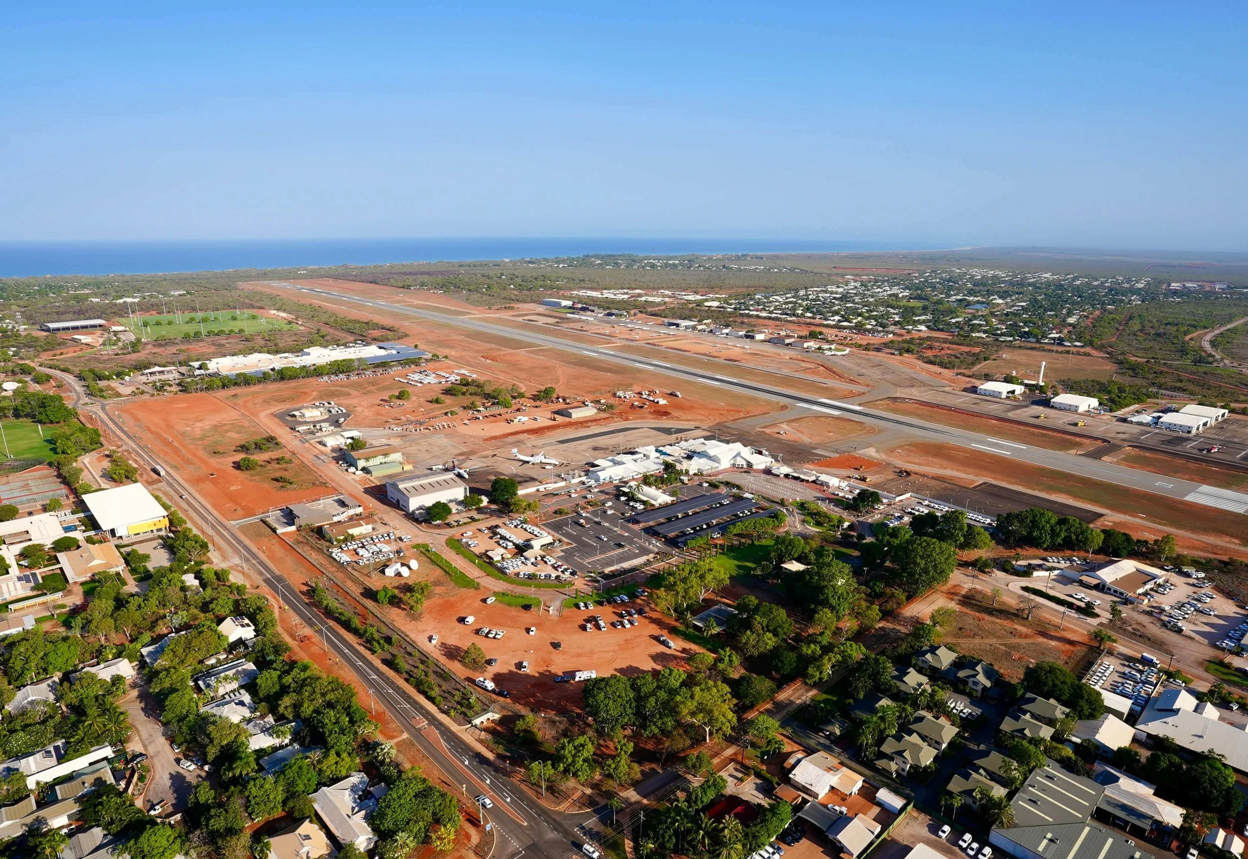 Aerial view of an airport runway with surrounding buildings, parking lots, residential houses, and a coastline in the distance under a clear blue sky.
