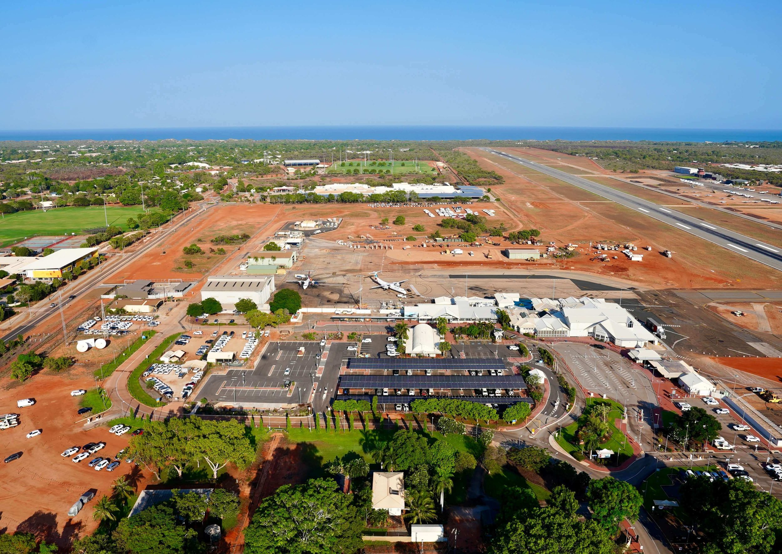 Aerial view of an airport with runways, airplanes, parking lots, green fields, and a distant ocean under a clear blue sky.