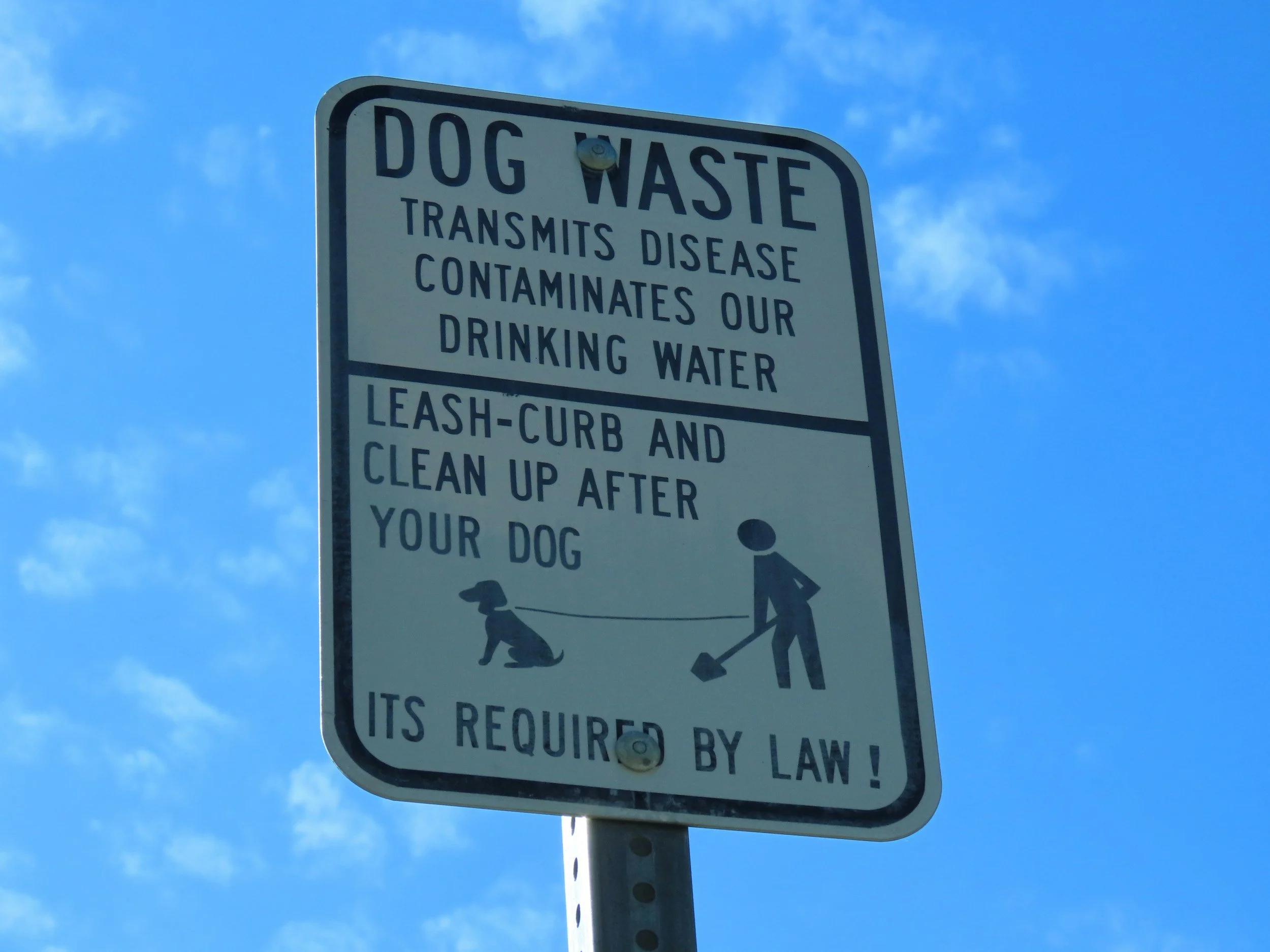 A sign indicating that dog waste transmits disease and contaminates drinking water, with illustrations of a person walking a dog and picking up waste.