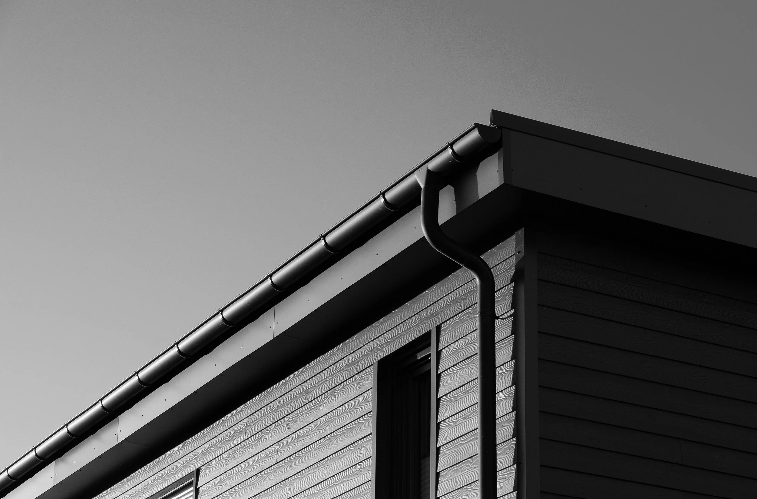 Black and white photo of a house's corner with a window, showcasing the roof, gutter, and siding.