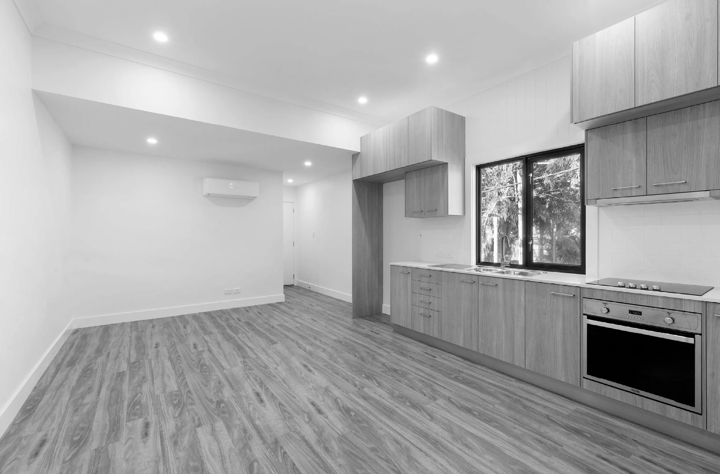 A bright, modern kitchen with wooden cabinets, a window overlooking trees, and an open living space with wood flooring.