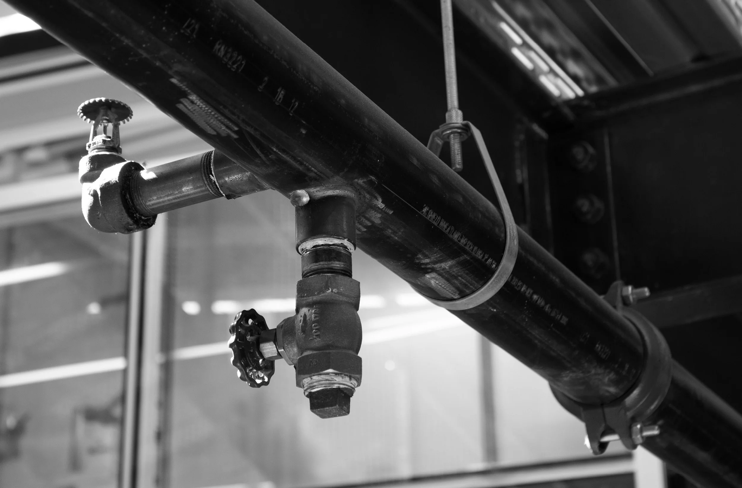 Black and white photo of a pipeline with a valve and a hanging cleanout pipe fitting, secured by metal brackets.