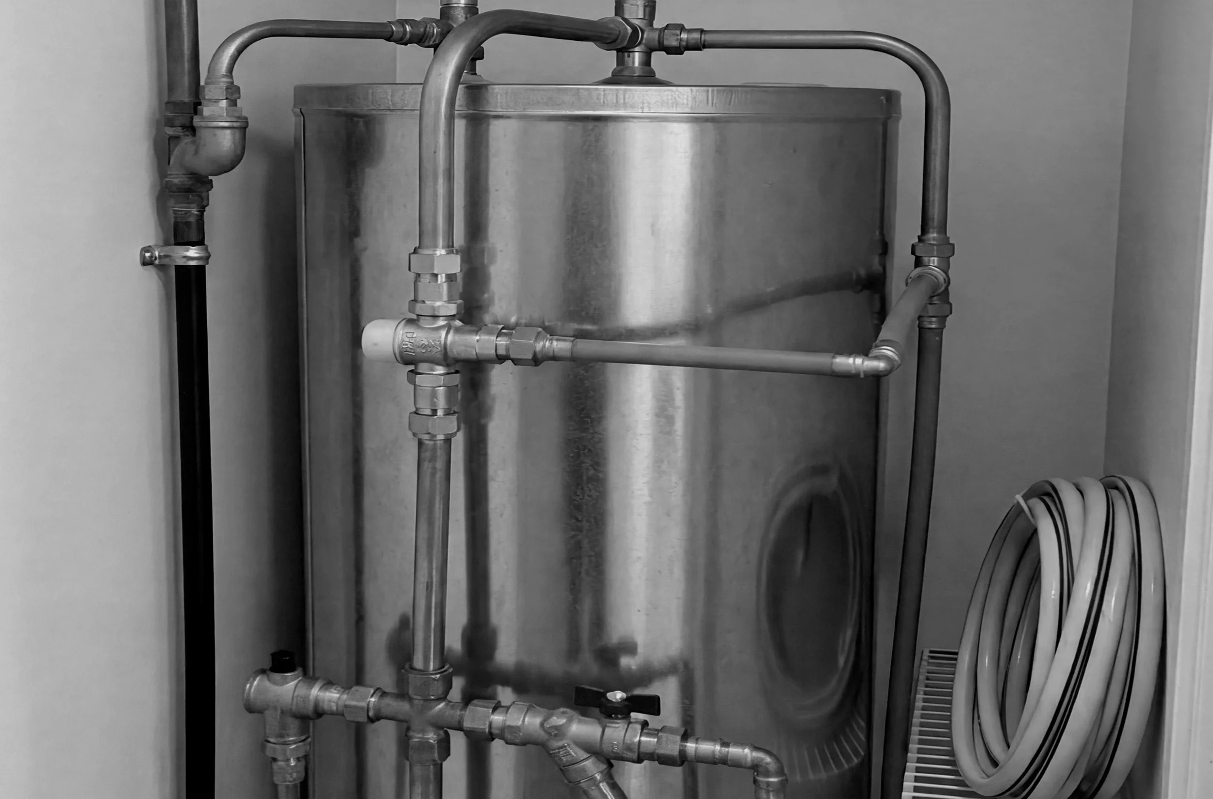 A large stainless steel water tank with several metal pipes connected to it, mounted in a corner of a room with plain walls.