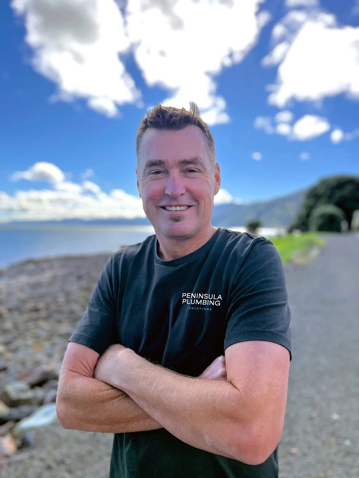 A smiling man with crossed arms standing outdoors on a rocky beach with water, mountains, and a partly cloudy sky in the background, wearing a black t-shirt with "PENINSULA PLUMBING SOLUTIONS" written on it.
