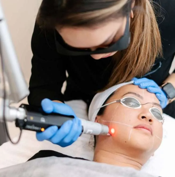 A person receives a facial skincare treatment using a handheld device, while lying down with protective eyewear, a headband, and a white towel on their head, administered by a professional wearing blue gloves.