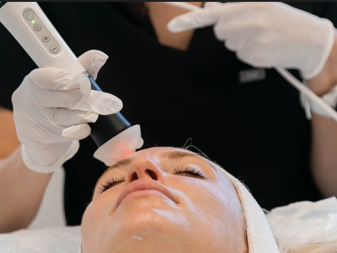 A person receiving a facial treatment from a professional using a handheld device while lying down with eyes closed.