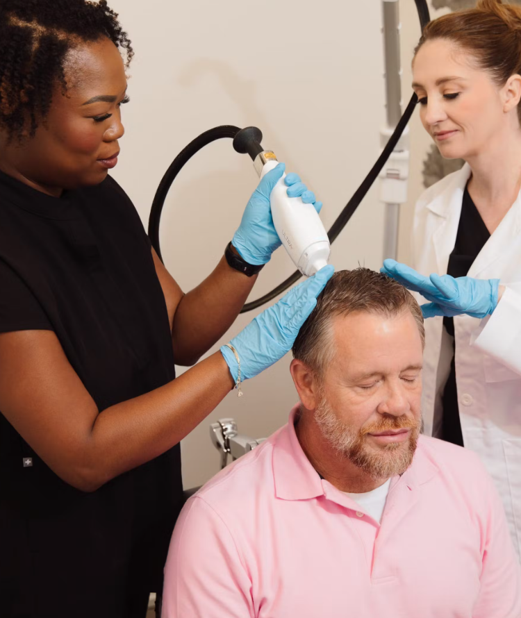 A healthcare professional performs a scalp treatment on a middle-aged man with gray hair and a beard, with another healthcare worker observing.