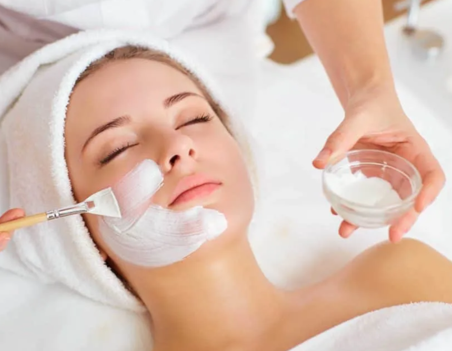 A woman with a towel wrapped around her head receives a facial treatment at a spa. She has her eyes closed as a skincare product is being applied to her face with a brush, while another person holds a bowl of cream or mask.