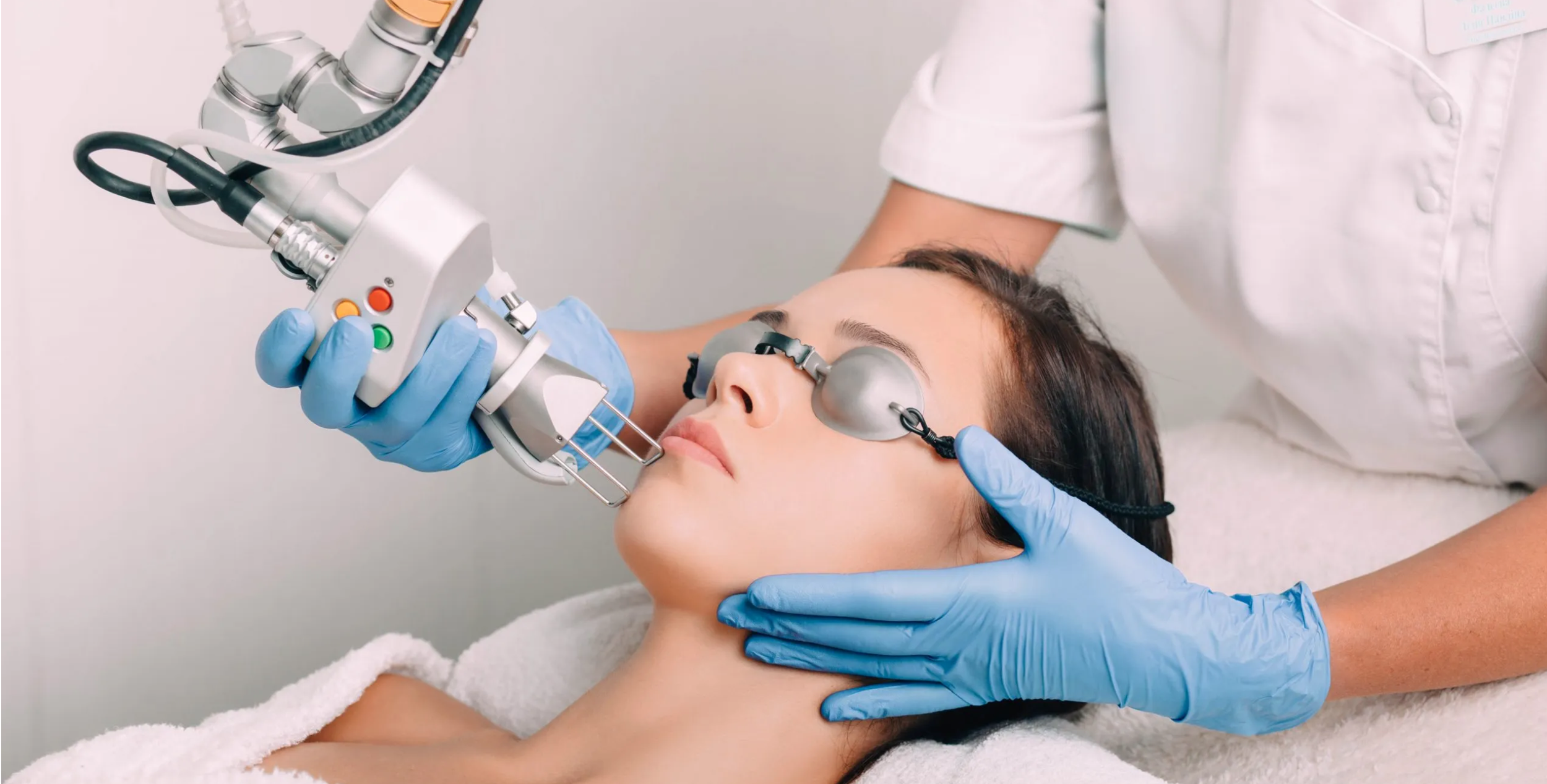 A woman receiving a cosmetic or dermatological treatment with a robotic device in a clinical setting, wearing protective goggles, with a healthcare professional attending to her.